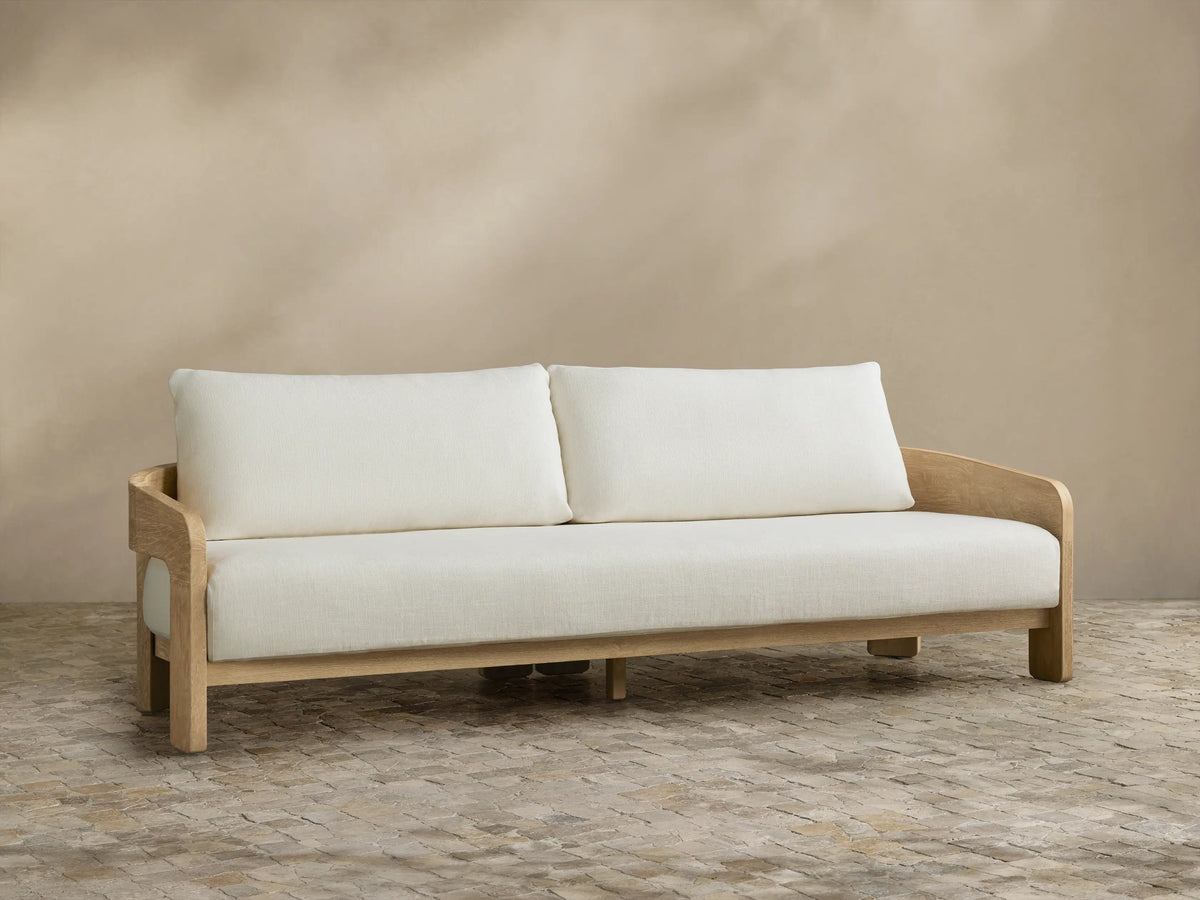 Anami Outdoor Sofa with light teak frame and two off-white seat & back cushions, on stone pavers against a beige wall.