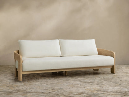 Anami Outdoor Sofa with light teak frame and two off-white seat & back cushions, on stone pavers against a beige wall.