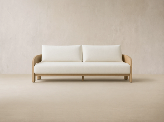 Anami Outdoor Sofa with light teak frame and two off-white seat & back cushions, on stone pavers against a beige wall.