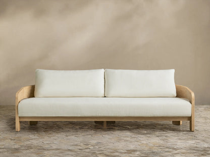 Anami Outdoor Sofa with light teak frame and two off-white seat & back cushions, on stone pavers against a beige wall.