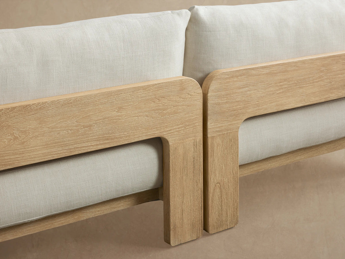 Anami Outdoor Sofa, light natural teak wood frame with two cream seat cushions, front view against a light brown wall.