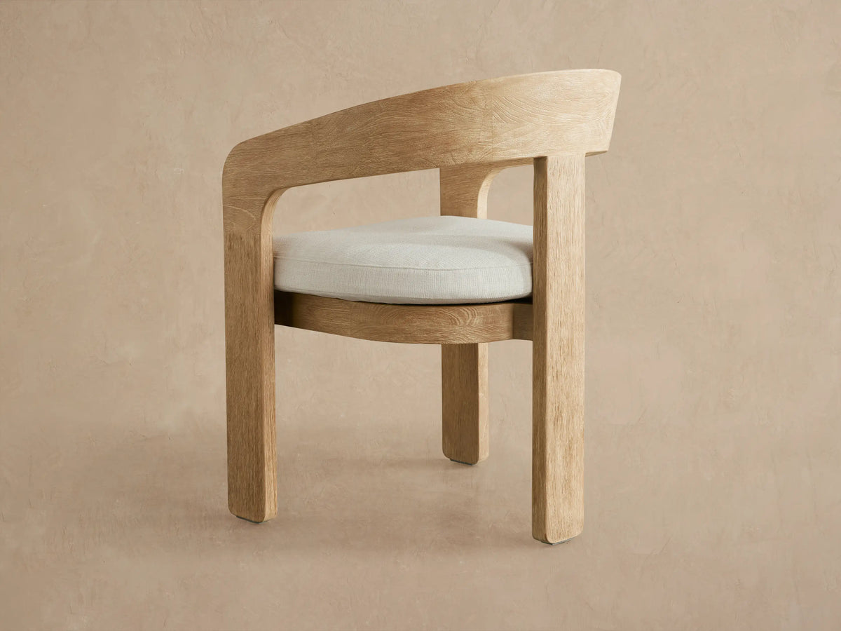 Close-up of Anami Outdoor Dining Chair: curved light teak wood backrest with prominent grain, off-white cushion, angled view.