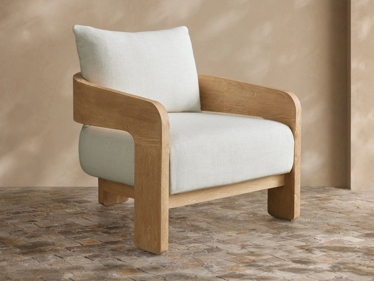 Anami Outdoor Lounge Chair with a light teak wood frame and plush off-white cushions on a patterned tiled floor.