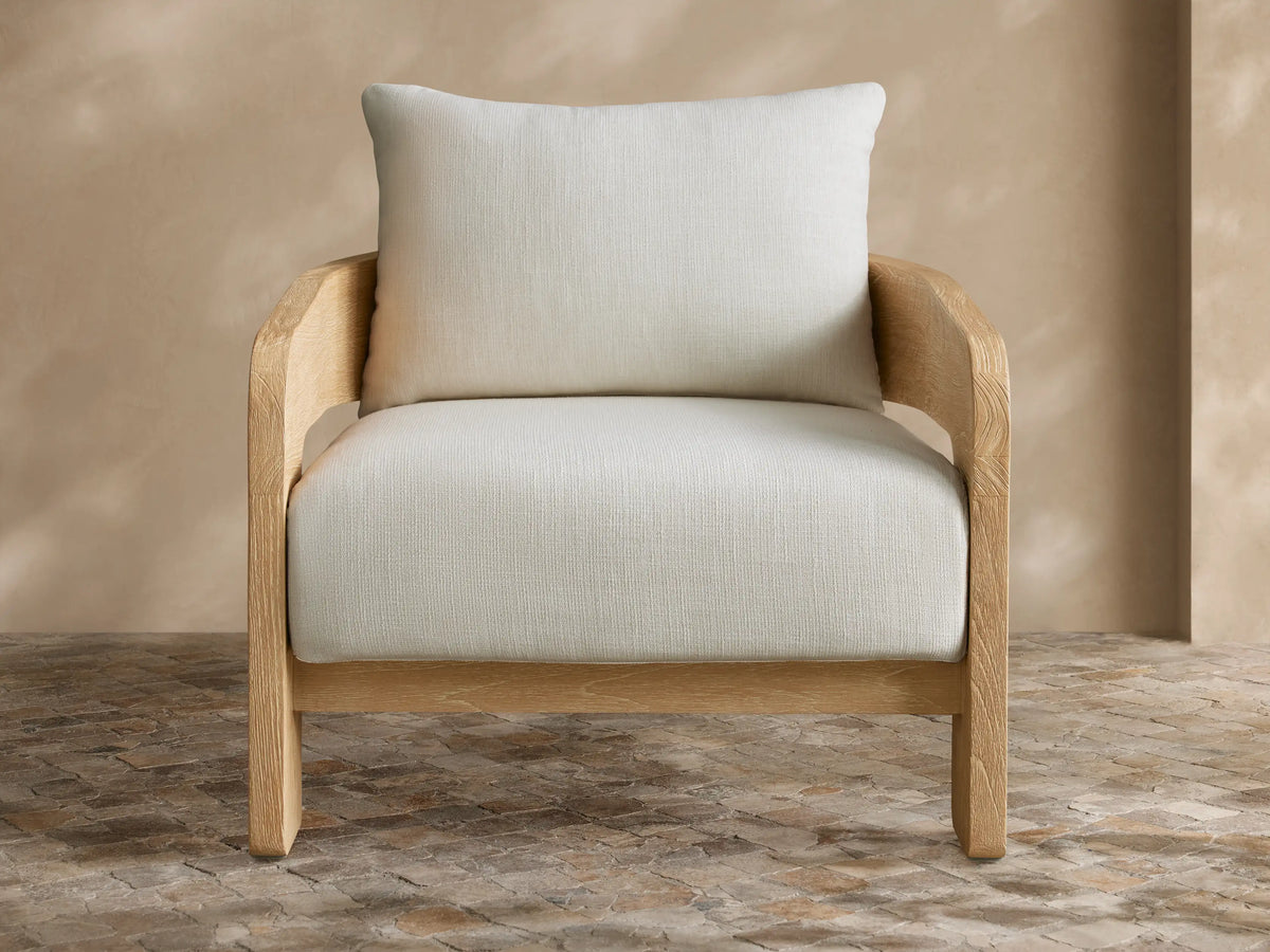 Anami Outdoor Lounge Chair with a light teak wood frame and plush off-white cushions on a patterned tiled floor.
