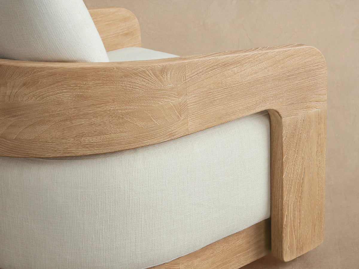 Anami Outdoor Swivel Chair with light wood frame and cream cushions, head-on view on stone floor and beige wall.
