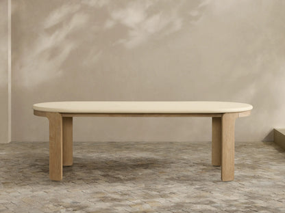 Anami Outdoor Dining Table with creamy oval top & sturdy, rectangular teak wood legs, against a beige wall with shadows.