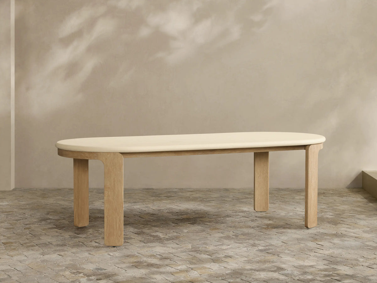 Anami Outdoor Dining Table with creamy oval top & sturdy, rectangular teak wood legs, against a beige wall with shadows.