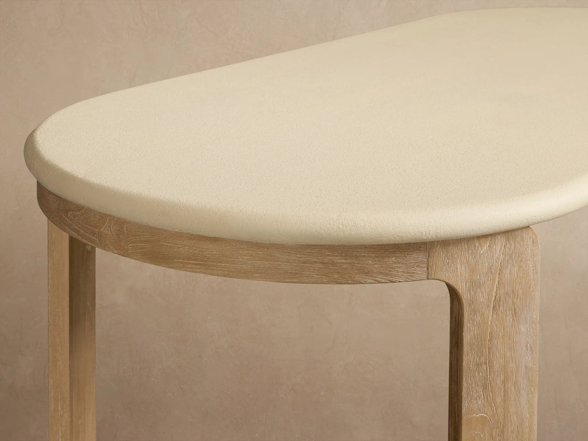 Anami Outdoor Dining Table with light cream oval top and natural teak wood legs on a textured stone floor, outdoors.