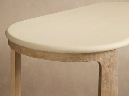 Anami Outdoor Dining Table with light cream oval top and natural teak wood legs on a textured stone floor, outdoors.