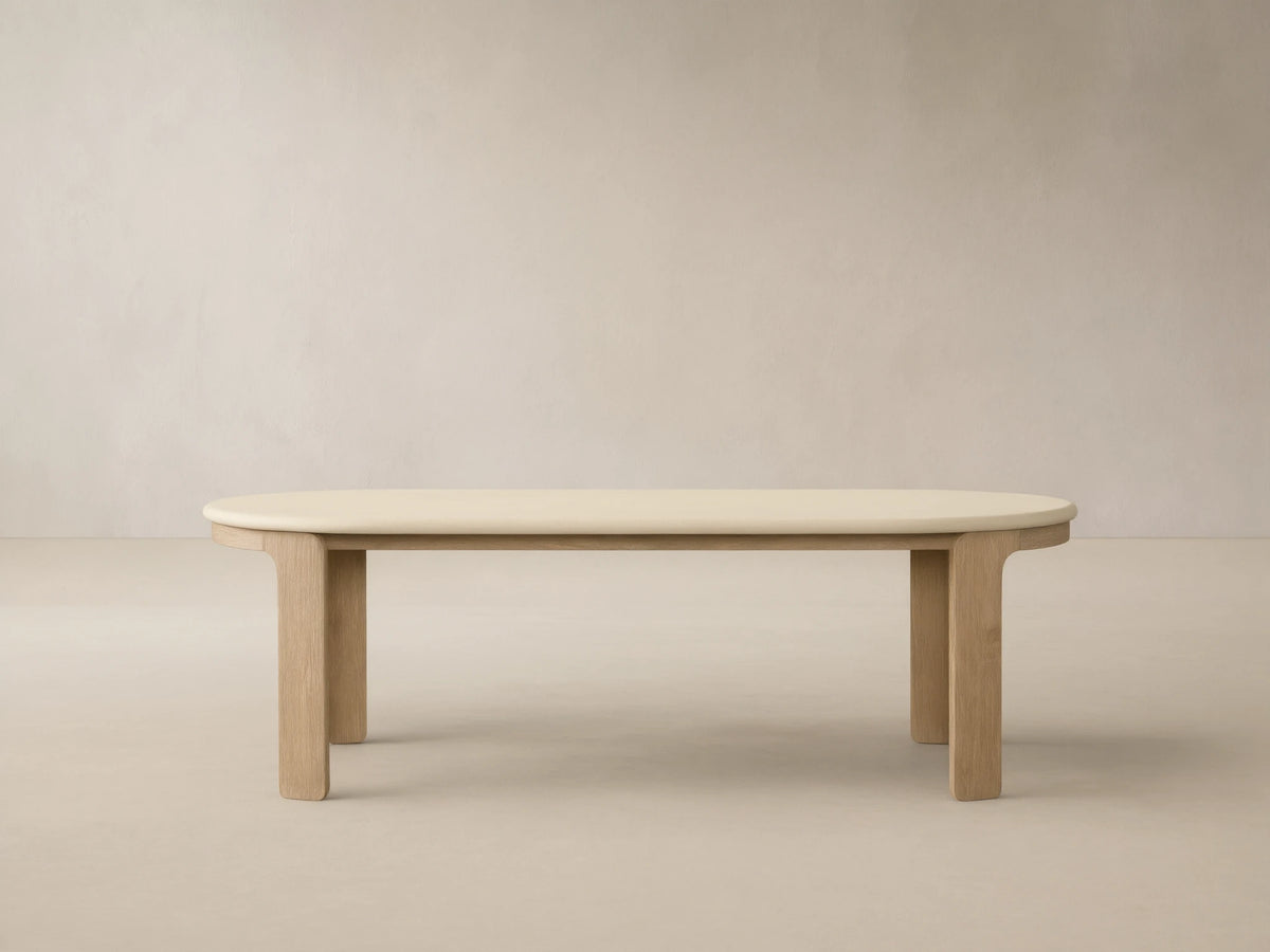 Anami Outdoor Dining Table with creamy oval top & sturdy, rectangular teak wood legs, against a beige wall with shadows.