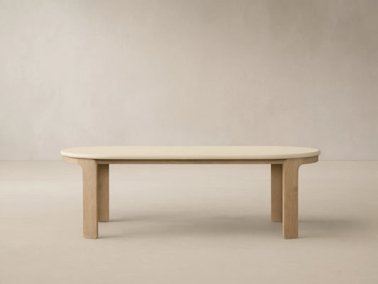 Anami Outdoor Dining Table with creamy oval top & sturdy, rectangular teak wood legs, against a beige wall with shadows.