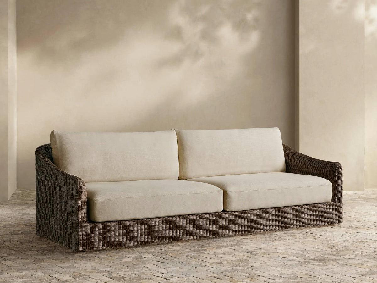 Ancona Outdoor Sofa, front view. Dark brown handwoven wicker frame with plush cream cushions on a patio with a tan wall.