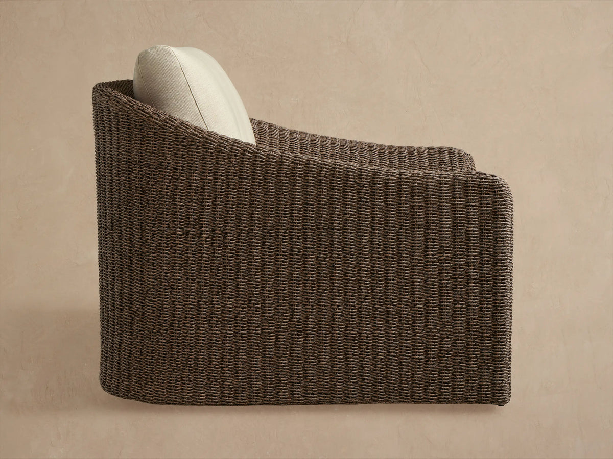 Close-up of Ancona Outdoor Swivel Chair, highlighting dark brown woven wicker armrest & light beige back/seat cushions.