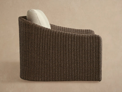 Close-up of Ancona Outdoor Swivel Chair, highlighting dark brown woven wicker armrest & light beige back/seat cushions.