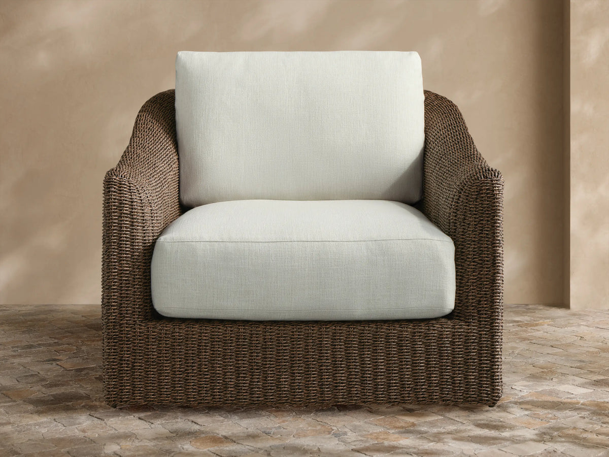Light brown woven Ancona Outdoor Swivel Chair with cream cushions, striped pillow, and side table in a garden patio.