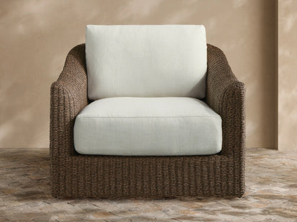 Light brown woven Ancona Outdoor Swivel Chair with cream cushions, striped pillow, and side table in a garden patio.