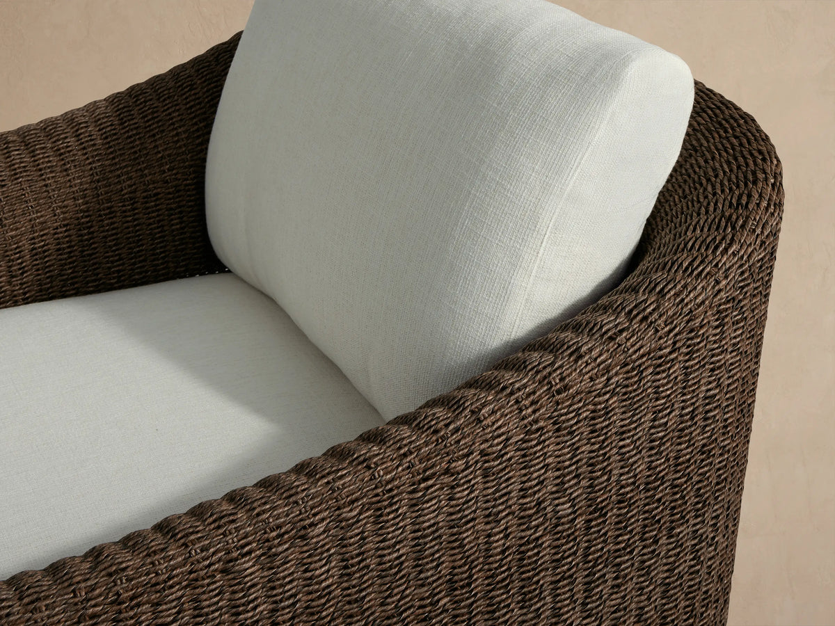 Ancona Outdoor Swivel Chair, front view, dark brown woven wicker frame & light cream cushions on a stone patio.