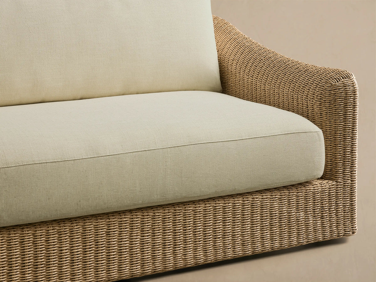 Front view of the Ancona Outdoor Sofa with light cream cushions and a tan, handwoven wicker base in an outdoor setting.