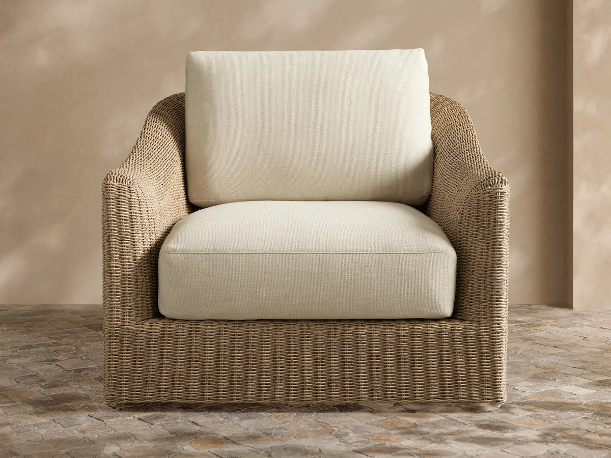 Natural wicker Ancona Outdoor Swivel Chair™ with light beige cushions and a striped pillow on an outdoor patio.