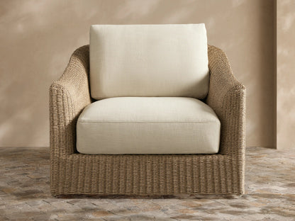 Natural wicker Ancona Outdoor Swivel Chair™ with light beige cushions and a striped pillow on an outdoor patio.