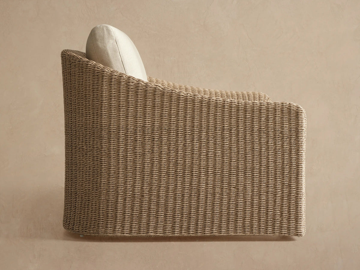 Close-up of Ancona Outdoor Swivel Chair with light tan handwoven wicker back and armrest, featuring a soft off-white cushion.