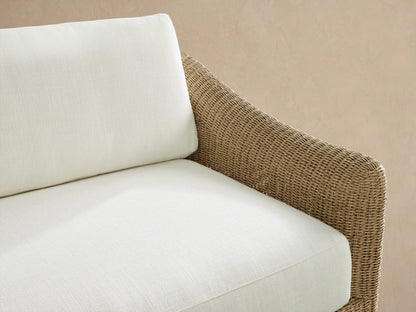 Ancona Outdoor Sofa, a two-seater with tan woven wicker frame and crisp white cushions, on a tiled patio against a light wall.