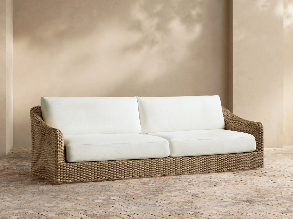 Ancona Outdoor Sofa: light brown woven wicker frame, two off-white cushions. Set against a warm beige wall with sunlight.