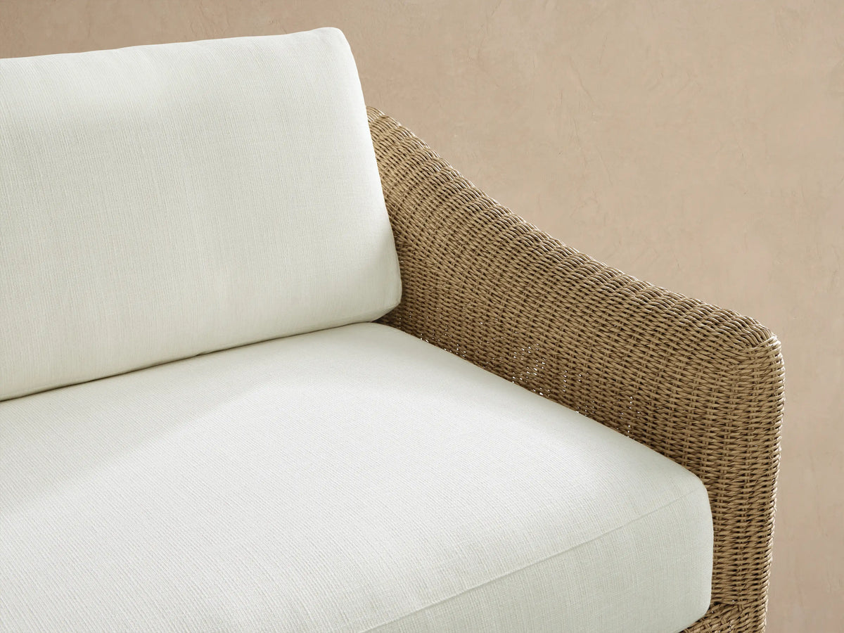 Ancona Outdoor Sofa, a 2-seater with a natural woven wicker frame and white cushions, on a stone patio.