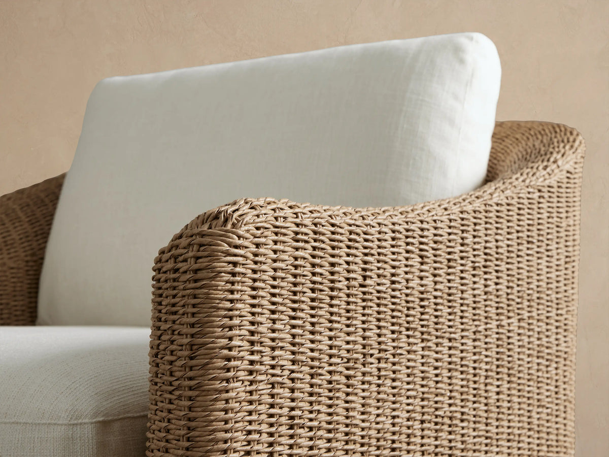 Ancona Outdoor Swivel Chair, front view. Light brown woven wicker chair with thick off-white seat and back cushions on a patio.