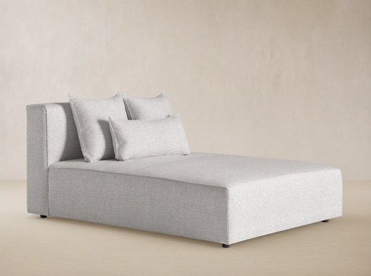Light gray textured Andaz Outdoor Chaise, modern, blocky design, with two square and one kidney pillow, against a beige wall.