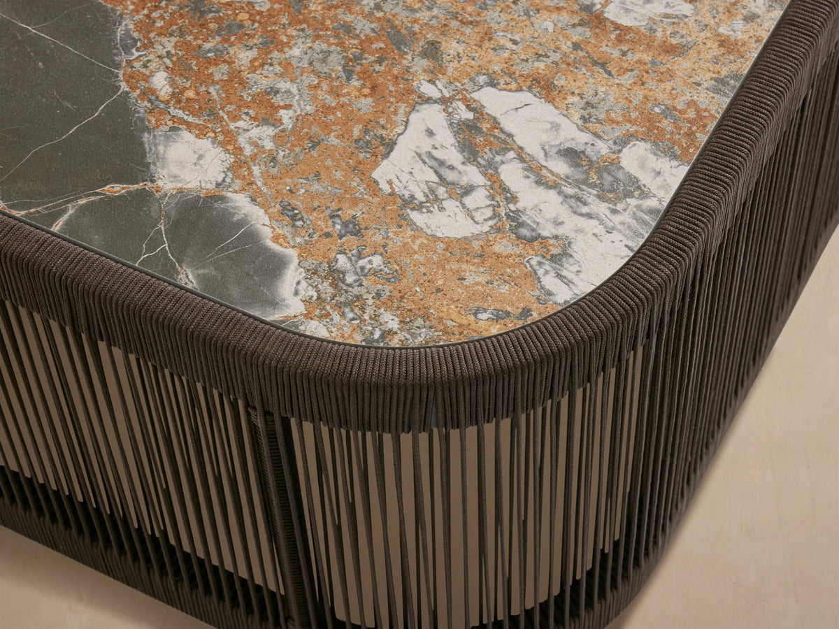 Anzio Outdoor Coffee Table. Oblong dark brown rope-wrapped base with linear pattern, mottled green/brown/white stone top. Outdoor patio scene.