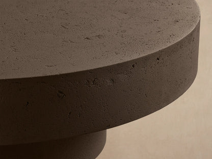 Dark brown, textured Azores Outdoor Round Coffee Table with a round top and cylindrical base, on a stone patio.