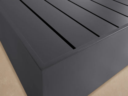 Bal Harbour Outdoor Aluminum Coffee Table: low, dark gray, rectangular, with a slatted top, on a rustic stone patio.