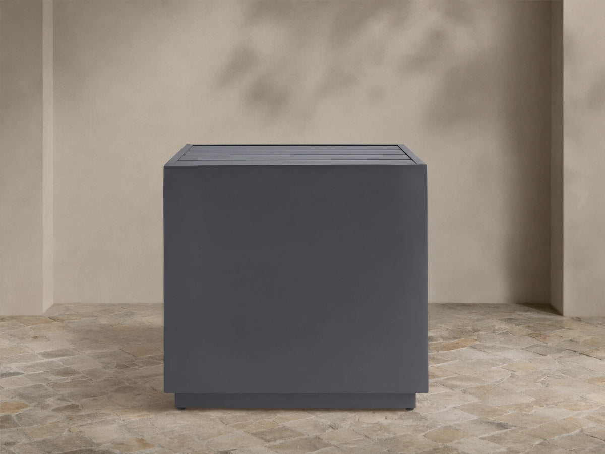 Dark gray Bal Harbour Outdoor Aluminum End Table, square with a slatted top, on a stone patio against a light wall.