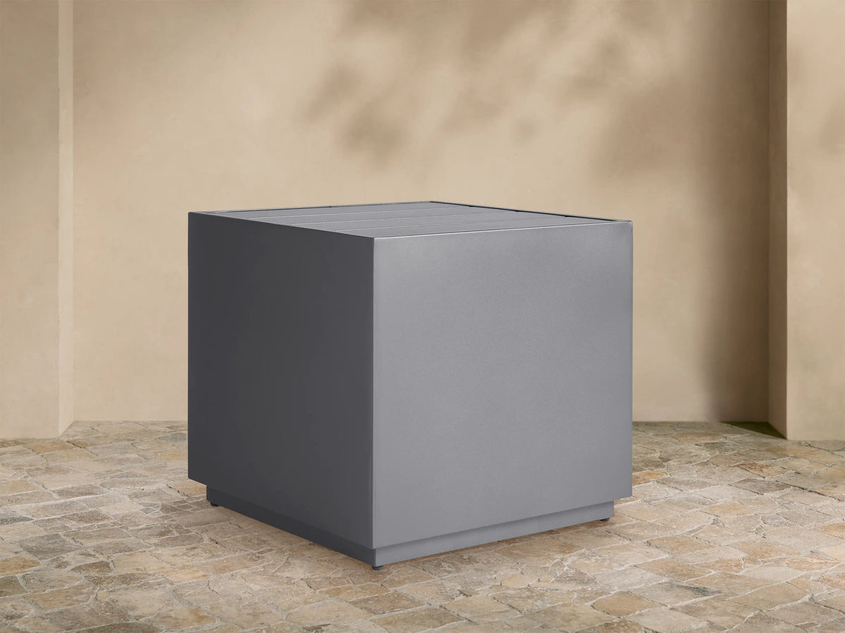 Dark gray Bal Harbour Outdoor Aluminum End Table, square with a slatted top, on a stone patio against a light wall.