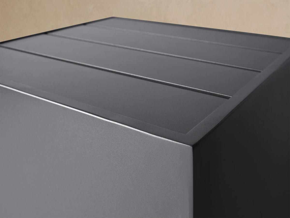 Dark grey cube-shaped Bal Harbour Outdoor Aluminum End Table with a slatted top, resting on a stone patio near a light wall.