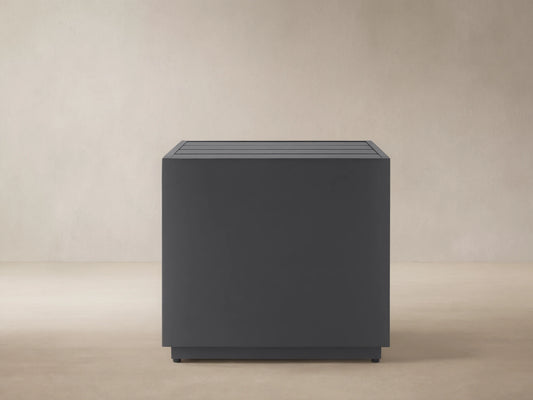 Dark gray Bal Harbour Outdoor Aluminum End Table, square with a slatted top, on a stone patio against a light wall.