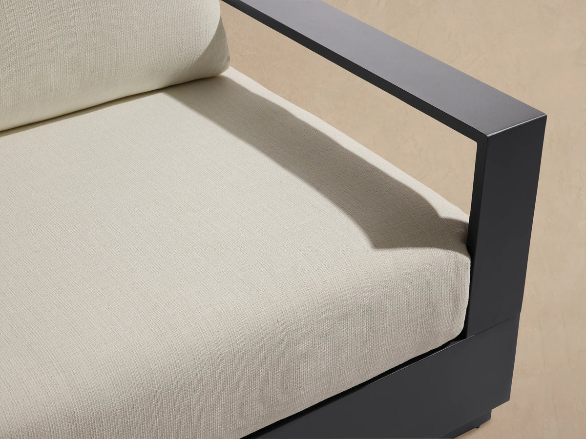 Bal Harbour Outdoor Aluminum Sofa, dark gray frame with light beige cushions, angled on a stone patio with a textured wall.
