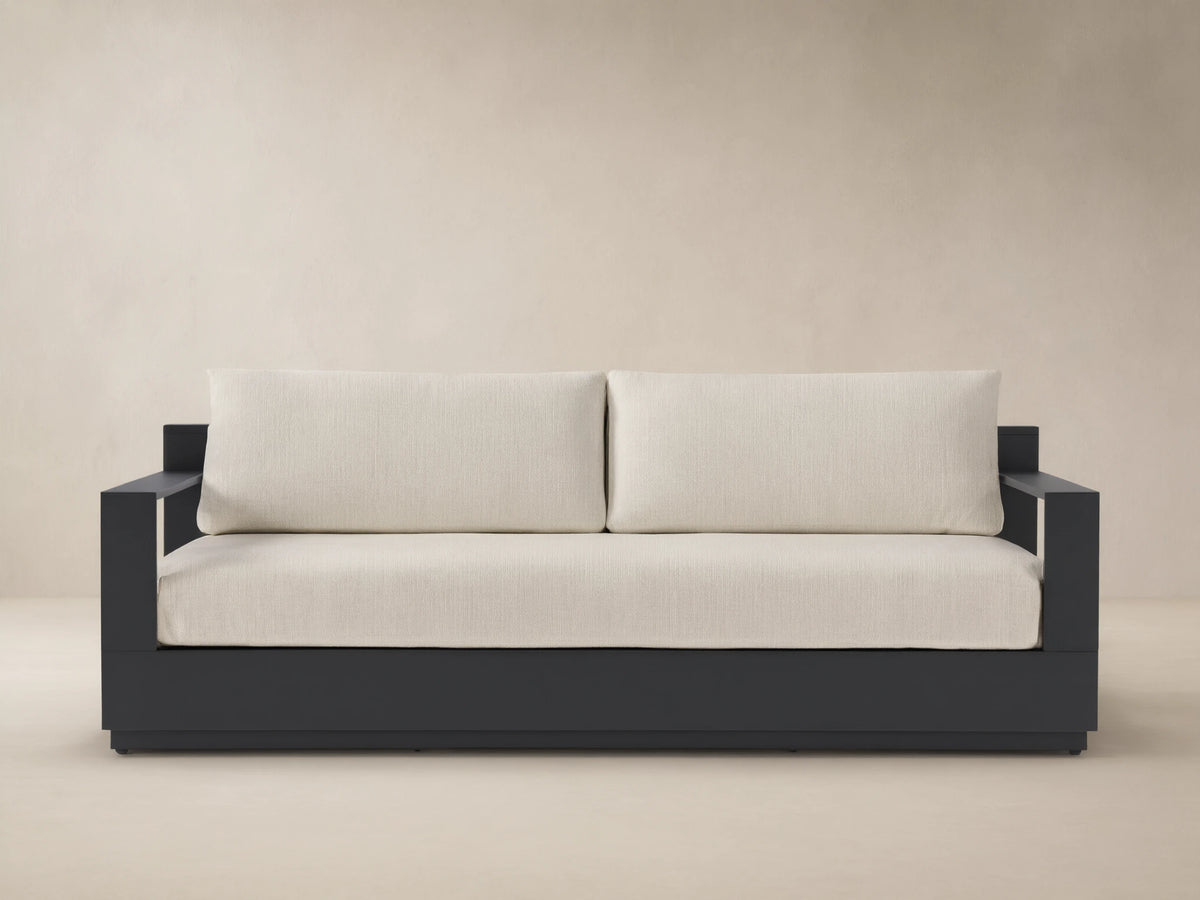Bal Harbour Outdoor Aluminum Sofa with dark gray frame and light beige cushions, centered on a patio against a neutral wall.