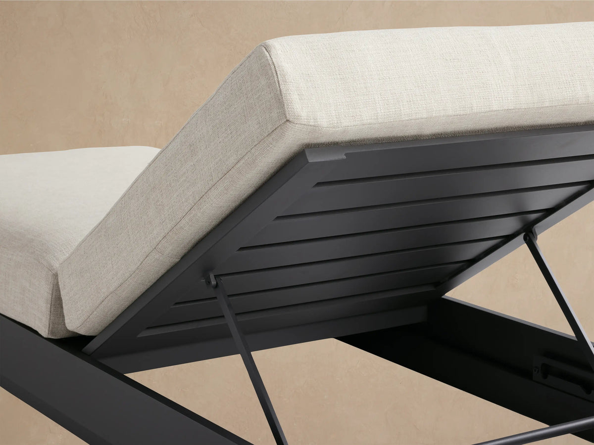 Bal Harbour Outdoor Aluminum Chaise with dark gray frame and light cream cushions. Backrest is elevated, side view against a warm tan wall.