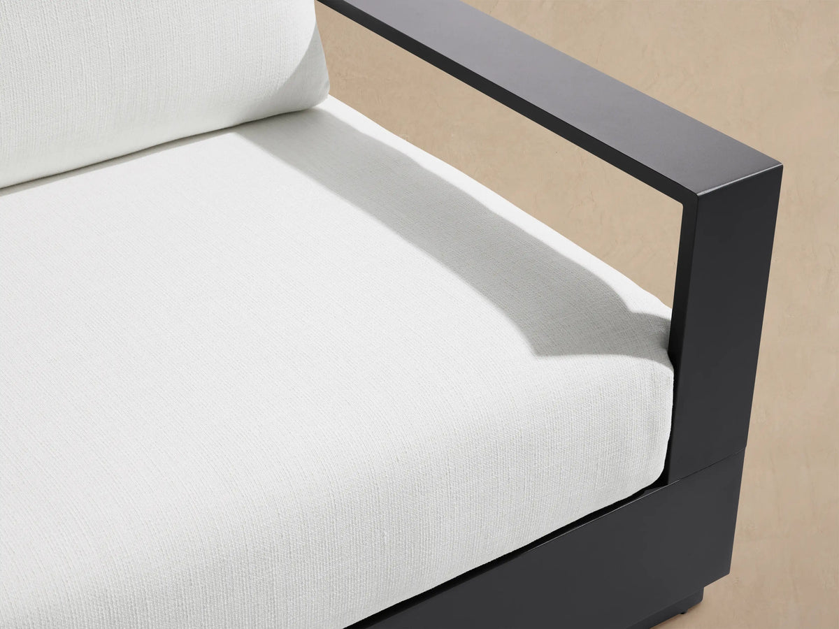Bal Harbour Outdoor Aluminum Sofa with a dark gray frame and white cushions, on a light tiled patio by a beige wall.