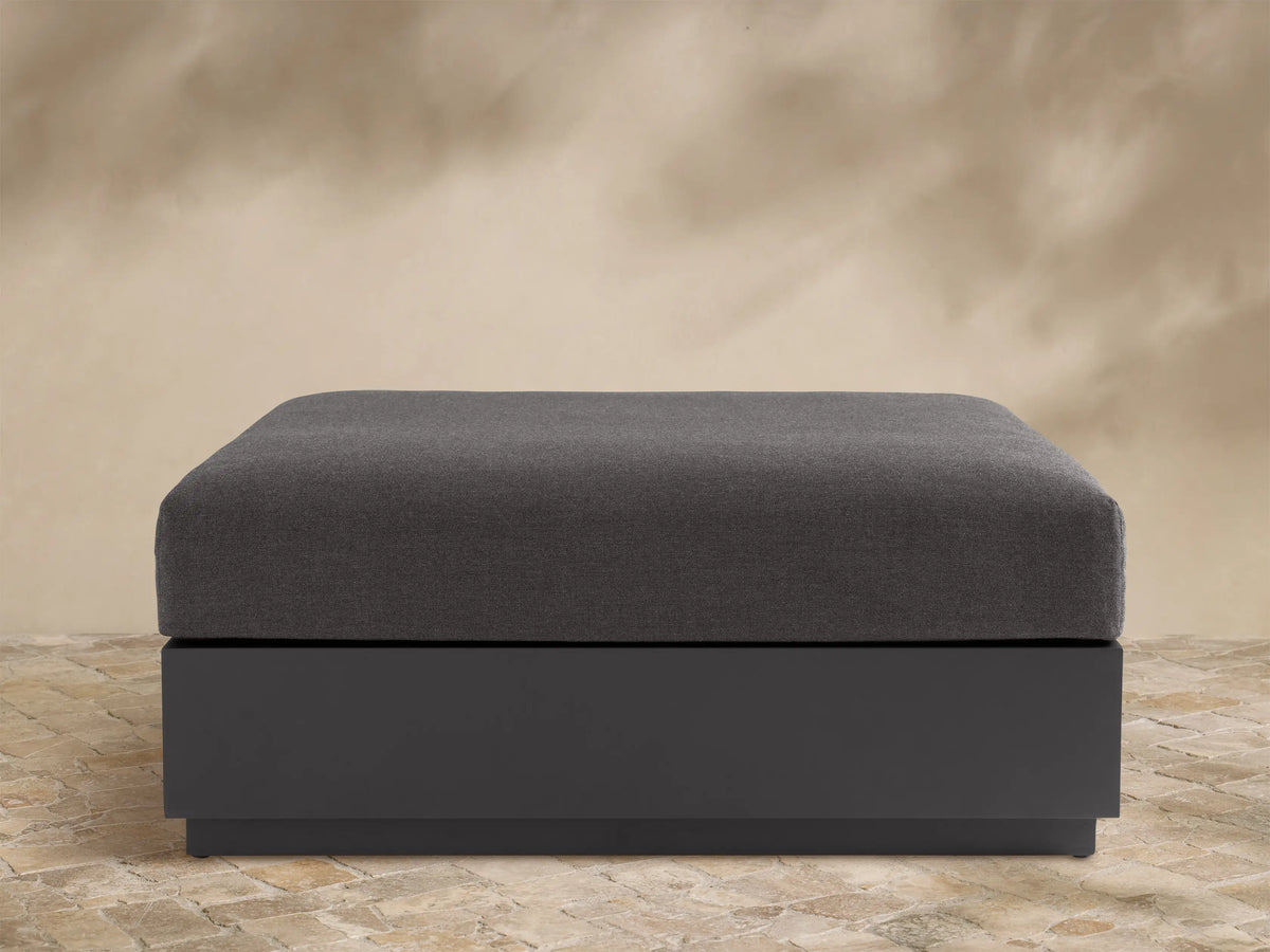 Bal Harbour Outdoor Aluminum Ottoman with a dark gray fabric cushion and a matte dark gray aluminum base on a stone floor.