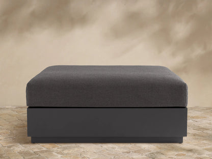 Bal Harbour Outdoor Aluminum Ottoman with a dark gray fabric cushion and a matte dark gray aluminum base on a stone floor.