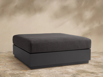Bal Harbour Outdoor Aluminum Ottoman with a dark gray fabric cushion and a matte dark gray aluminum base on a stone floor.