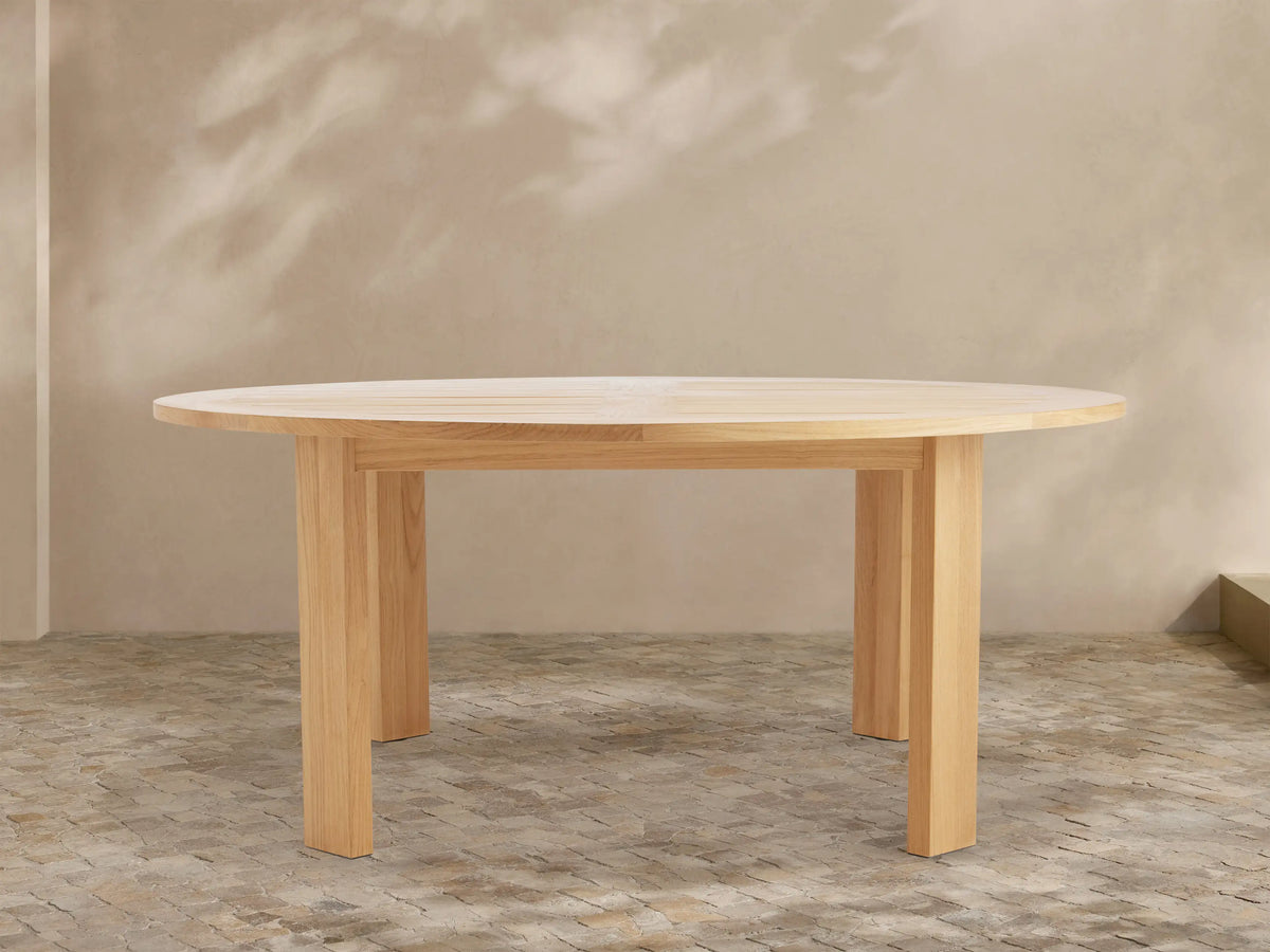 Round Bal Harbour outdoor dining table, light golden teak wood, with a slatted top and thick, sturdy legs on a tiled patio.