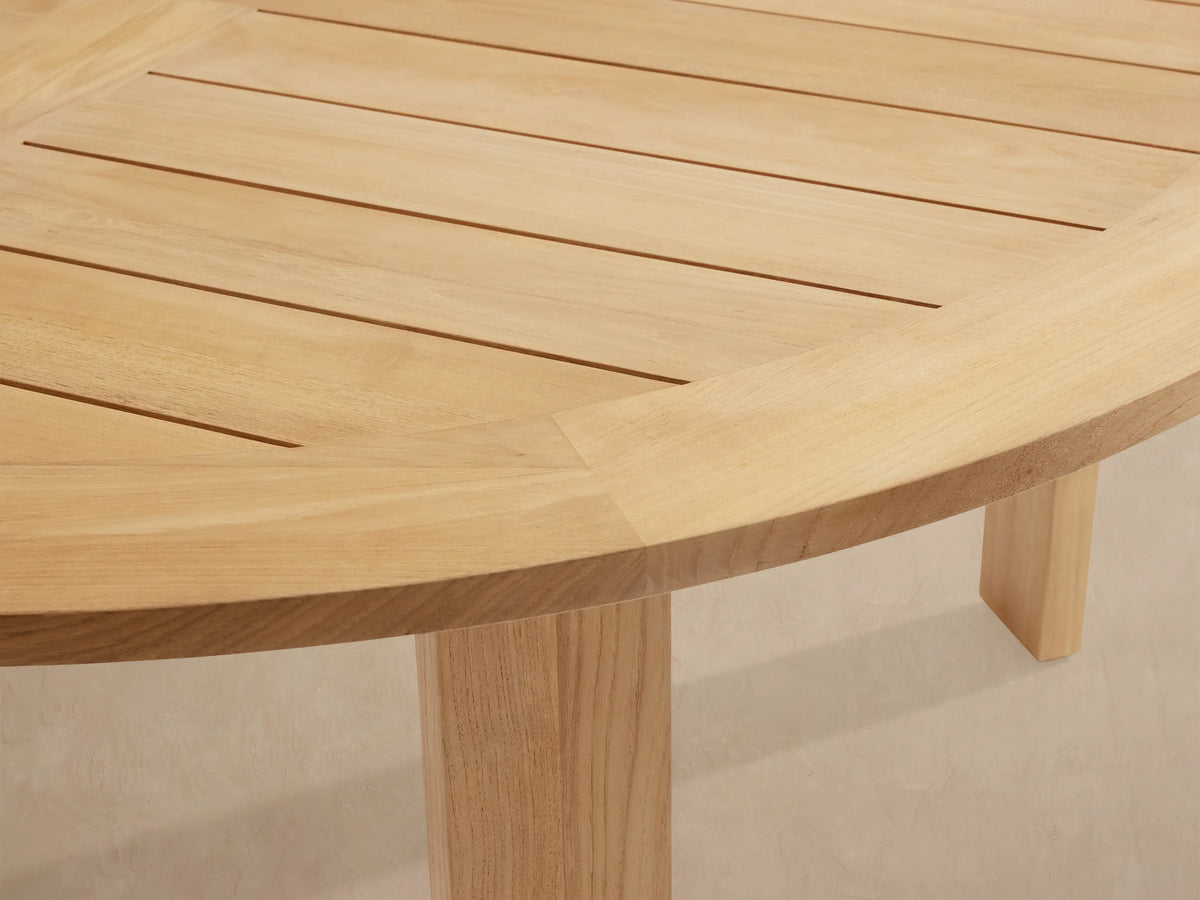 Round Bal Harbour outdoor dining table, light golden teak wood, with a slatted top and thick, sturdy legs on a tiled patio.