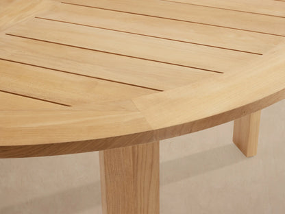 Round Bal Harbour outdoor dining table, light golden teak wood, with a slatted top and thick, sturdy legs on a tiled patio.