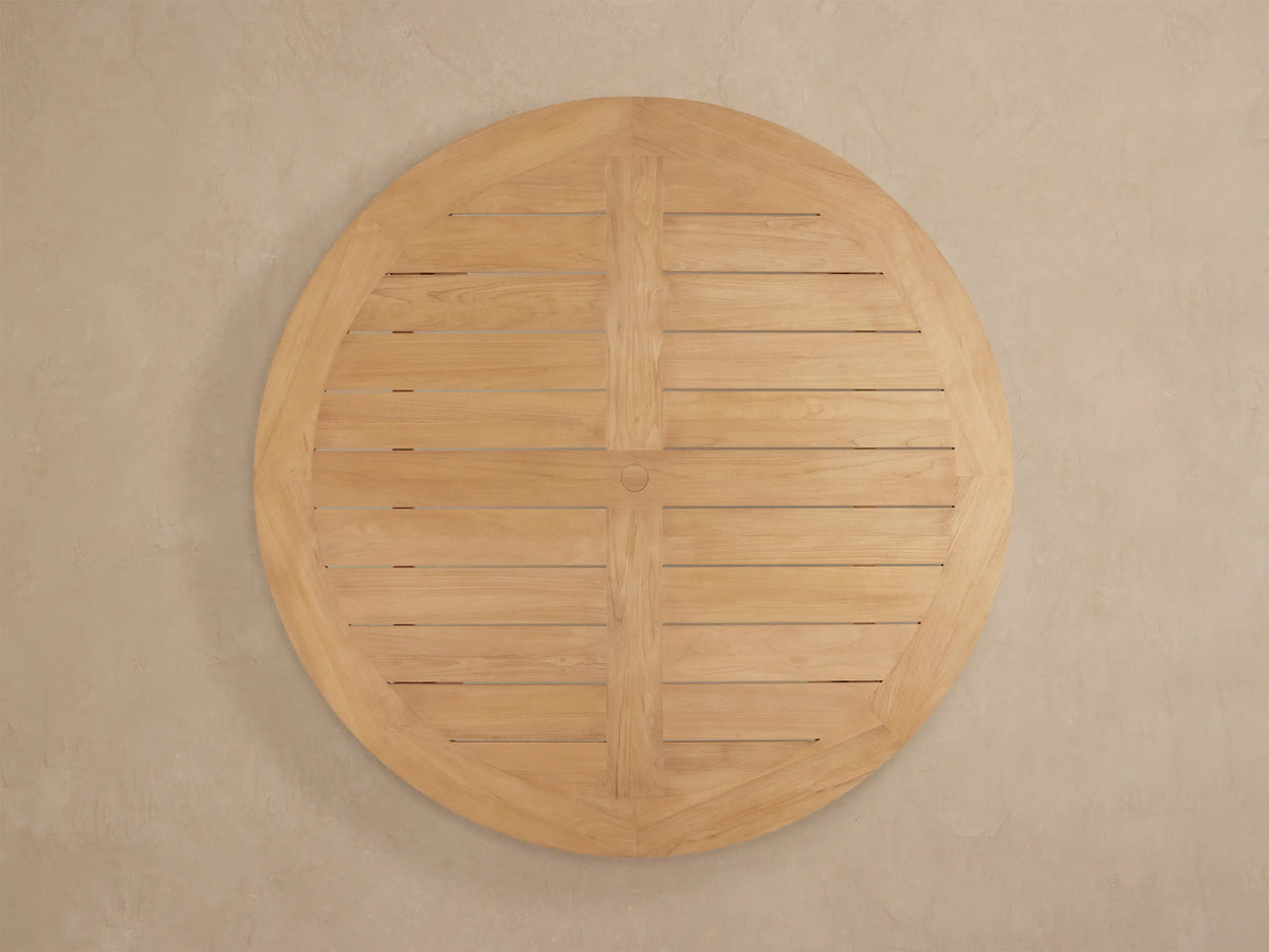 Close-up of Bal Harbour Outdoor Teak Round Dining Table; light golden-brown slatted top, natural wood grain, curved edge, sturdy leg.