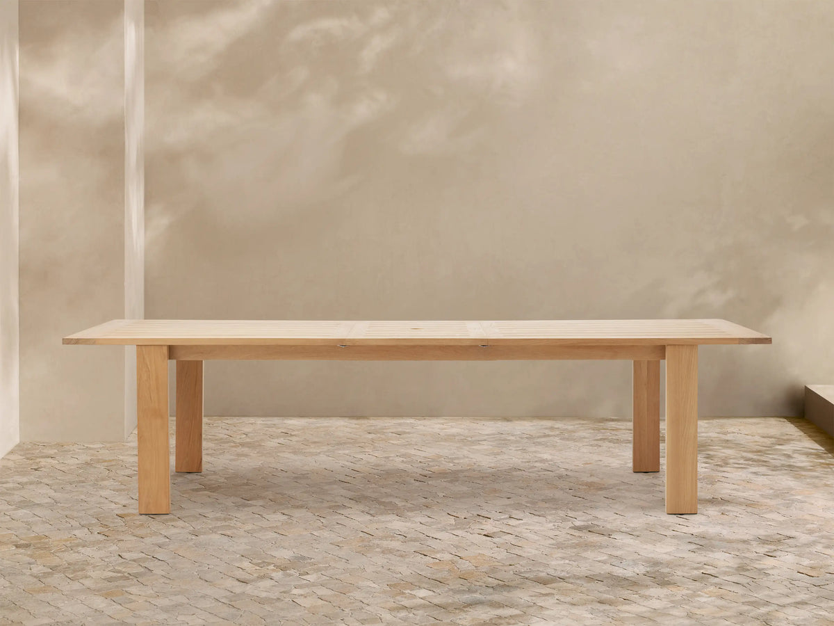 Bal Harbour Outdoor Teak Extension Dining Table, light teak wood with slatted top and sturdy legs, on a stone patio.