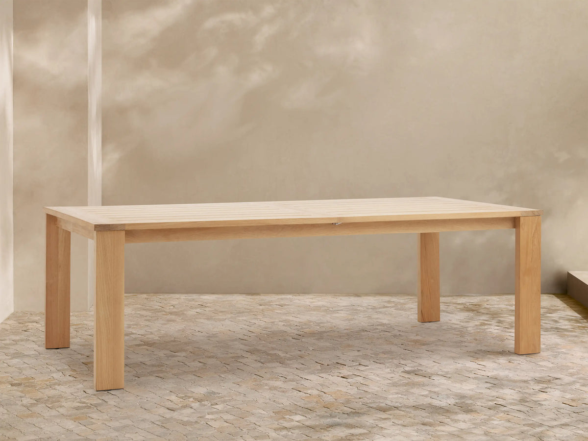 Bal Harbour Outdoor Teak Extension Dining Table, light natural wood, rectangular with slatted top, against a beige wall & stone floor.